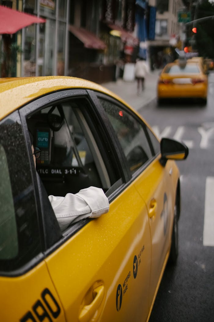 From above of unrecognizable person in white shirt sitting in modern yellow cab on asphalt roadway on urban background in town at daytime